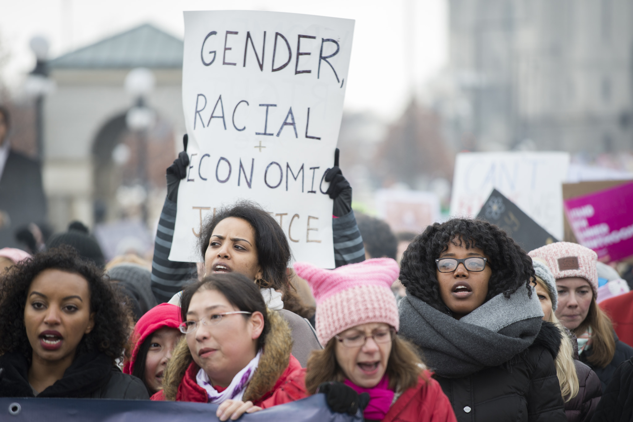 an image featuring a small group of protesters at a Women's March. The group is racially diverse and wearing warm winter clothes. Some of them wear pink pussy hats. One protester holds a sign that states Gender, Racial, Economic Justice