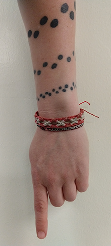 photogrpah of a white woman's arm with a tattoo of black spots circling up her arm. She is wearing three small, hemp wrist bands.
